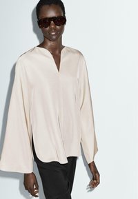Blouse beige, légère, avec un décolleté en V et des manches larges, dotée d'échancrures sur les côtés pour plus de mouvement, associée à un pantalon sombre et des lunettes de soleil oversize.