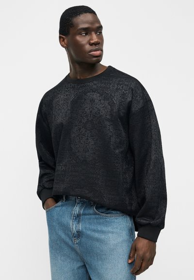 Άνδρας που φοράει μαύρο μακρυμάνικο φούτερ με σχέδια και μπλε τζιν παντελόνι, κοιτάζει προς τα πλάγια με το ένα χέρι στην τσέπη, με απλό φόντο.