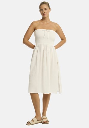 Mujer de pie con las manos detrás de la espalda, vistiendo un vestido midi blanco sin tirantes y sandalias beige deslizantes sobre un fondo liso y claro.