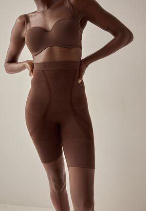 Žena u smeđim stezajućim kratkim hlačama visokog struka i pripadajućem grudnjaku, stoji s rukama na bokovima ispred jednobojne pozadine.