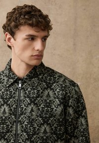 Dunkelgrüne Jacke mit kompliziertem floralen Muster in Hellgrün. Verfügt über einen Reißverschluss und einen Kragen. Der Stoff wirkt strukturiert und texturiert.