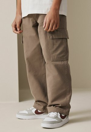 Persoon die losse bruine cargobroek en witte sneakers met bordeauxrode accenten draagt, staand op een neutrale vloer tegen een effen muur.