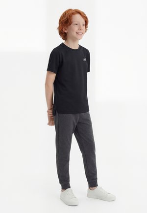 Rødhåret dreng, stående, iført sort kortærmet skjorte, grå joggebukser og hvide sneakers, smilende og kiggede til siden.