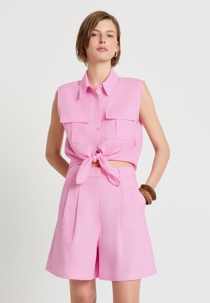 Femme portant une chemise rose sans manches à col, nouée à la taille, et un short taille haute assorti, avec des bracelets en bois au poignet, debout devant un fond blanc.
