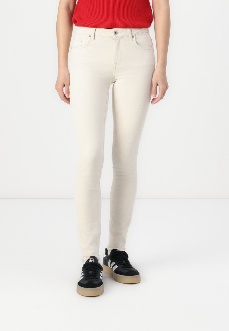 Jeans skinny beige con una superficie denim liscia, vita alta e design a cinque tasche, abbinati a sneakers nere con strisce bianche.