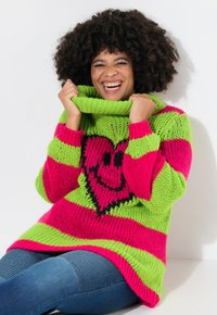 Pull en tricot coloré avec des rayures rose vif et vert, un grand motif en forme de cœur noir et un col montant haut.
