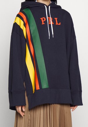 Marineblauer Hoodie mit weißen Kordeln, orangefarbenen "PRL"-Buchstaben und gelben, orangefarbenen und grünen diagonalen Streifen auf der linken Seite, getragen mit einem beigen Rock.