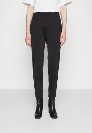 Vrouw draagt zwarte, op maat gemaakte broek tot op de enkel, zwarte laarzen met hak en een wit topje met korte mouwen, staand tegen een effen achtergrond.