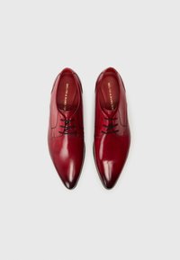 Červené kožené boty na šněrování s špičatou špičkou, minimálním prošíváním a designem na šněrování. Uvnitř se nachází značka ve zlaté barvě. Hladký povrch.