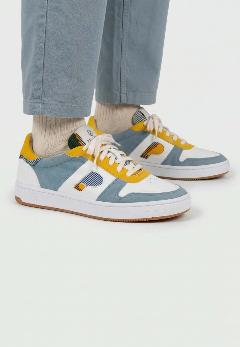 Pieds portant des baskets blanches, jaunes et bleues avec des chaussettes beiges et un pantalon bleu clair sur un fond blanc uni.