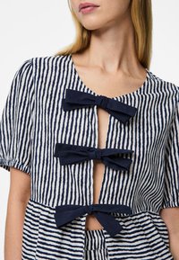 Blusa a righe in crema e blu navy con tre fiocchi in tessuto, maniche corte a palloncino e scollatura a V con chiusura a bottone.