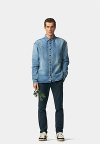Chemise en denim bleu clair avec un design à boutons, poche poitrine et manches longues ; associée à un pantalon sombre et des baskets, tenant une plante.