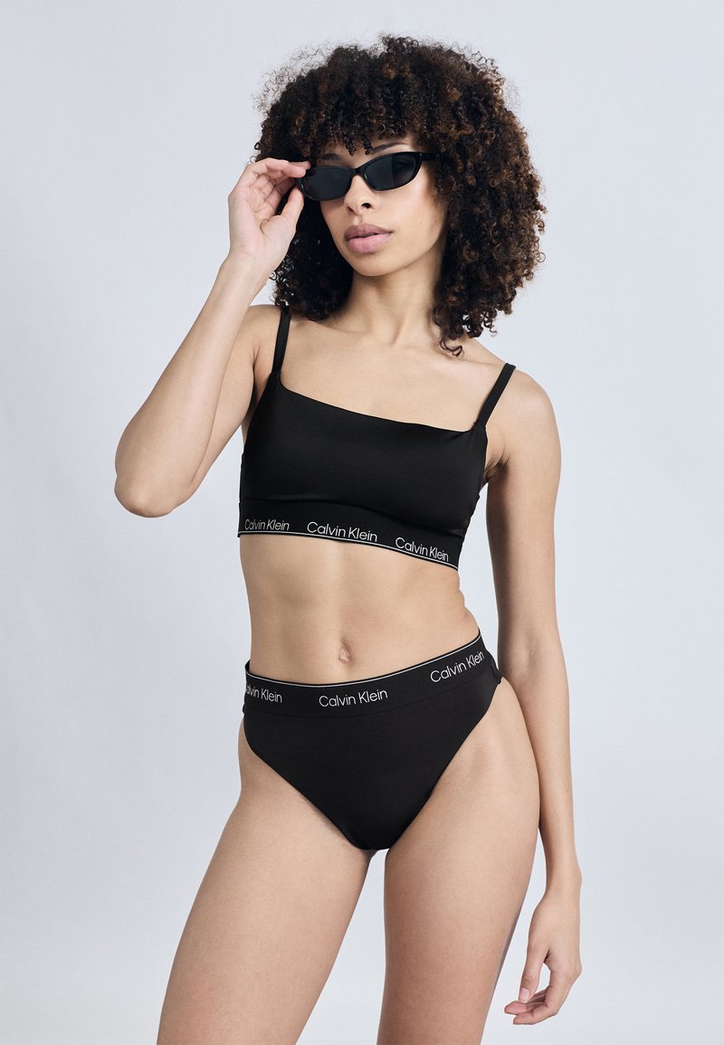 Μαύρο μπραλέτα με λεπτές τιράντες και λωρίδα με λογότυπο, σε συνδυασμό με μαύρο μπικίνι ψηλόμεσο που έχει αντίστοιχη λωρίδα μέσης με λογότυπο.