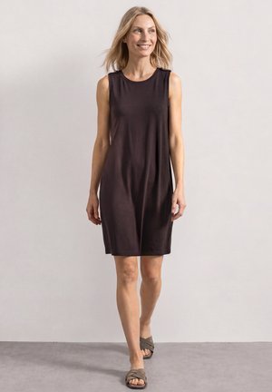 Frau, die ein ärmelloses, dunkelbraunes, knielanges Kleid und graue Sandalen trägt, vor einem schlichten hellen Hintergrund.