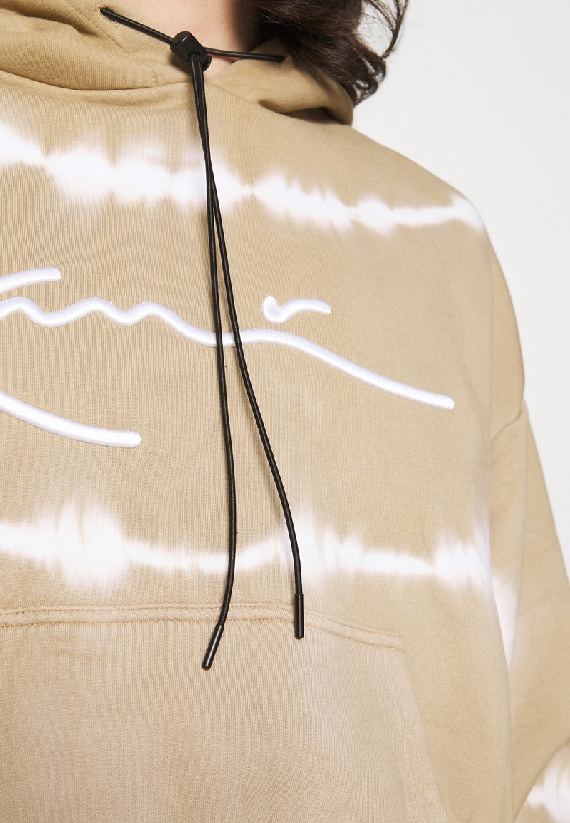 Sudadera beige con patrón de tie-dye en blanco. Presenta un bolsillo frontal, capucha con cordón y detalles en negro, y una textura de algodón suave.