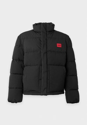 Jeune homme portant une doudoune noire, un pull gris zippé, un jean bleu et des baskets blanches, debout devant un fond blanc uni.