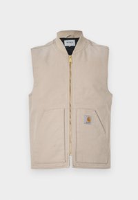 Beige vest med høy krage, frontlukking med glidelås, to store lommer og et lite logo-patch på venstre side. Laget av slitesterkt stoff.