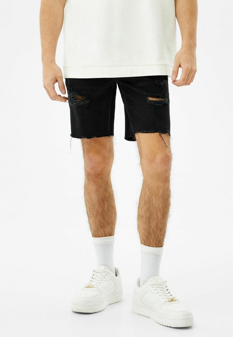 Černé džíny šortky s roztrhanými detaily a třepenými okraji, zkombinované s bílými teniskami a sportovními ponožkami, nošeny s oversize bílým topem.