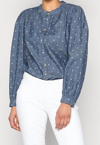 Lehká modrá bavlněná blůza s dlouhými rukávy, kulatým límcem, předními knoflíky a bílými křížovými vzory po celém povrchu. V kombinaci s bílými kalhotami.