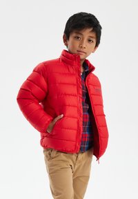 Giacca rossa in piuma con collo alto, design a righe orizzontali e tasche laterali. Indossata sopra una camicia a quadri blu e rossa.