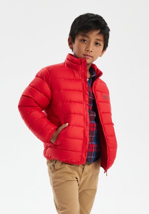 Giacca rossa in piuma con collo alto, design a righe orizzontali e tasche laterali. Indossata sopra una camicia a quadri blu e rossa.
