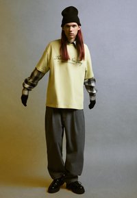 Tricou galben supradimensionat cu text grafic, purtat peste o cămașă în carouri negre și gri, asociat cu pantaloni lărgi gri și pantofi negri.