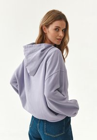 Felpa viola con cappuccio e vestibilità rilassata, caratterizzata da spalle abbassate e tessuto morbido, abbinata a jeans in denim blu.