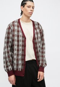 Cardigan en laine marron et grise duveteuse avec une bordure bordeaux, présentant un motif à carreaux et des boutons sur le devant, porté par-dessus une chemise beige.