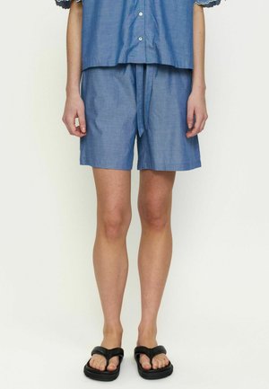 Személy kék gombos sortot és hozzá illő felsőt visel fekete szandálokkal, egyszerű világos háttér előtt áll.