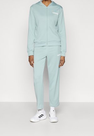 Person iført lyseblåt Adidas træningssæt med lynlås-hættetrøje og matchende bukser, hvide Adidas sneakers, stående på gråt gulv mod hvid væg.