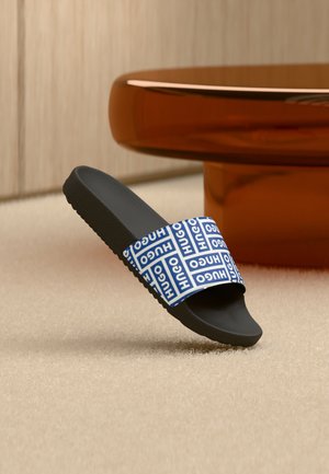 Schwarze Slidesandale mit blau-weißem HUGO-Logoband, die auf beigem Teppich neben einem runden, durchscheinenden braunen Tisch liegt.