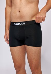 Man wearing black SNICKS brand boxer briefs, pulling waistband slightly away from body, showing torso and upper legs.
