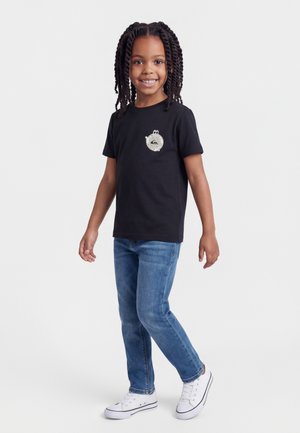 Naeratav laps põimitud juustega, kandes musta logoga T-särki, siniseid teksaseid ja valgeid tenniseid, seisab ühevärvilise tausta ees.