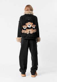 Chaqueta de mezclilla negra con cuello de piel, que presenta un diseño de oso de peluche en la parte de atrás. Combinada con jeans negros holgados y zapatos beige.