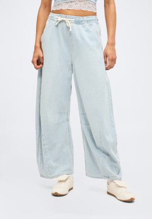 Άτομο που φοράει φαρδιά παντελόνια με ουροκόκκινες και λευκές ρίγες, με ζώνη με κορδόνι στη μέση, λευκά αθλητικά και ένα κοντό τοπ με δαντέλα.