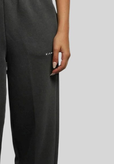 Mano destra con unghie beige che si appoggia accanto a pantaloni grigio scuro con piccolo testo bianco "RICH" sulla coscia contro uno sfondo grigio chiaro.