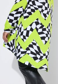 Persona che indossa una maglietta a maniche lunghe verde con un motivo a zigzag bianco e nero e un design a scacchi ondulato, abbinata a pantaloni in pelle nera.