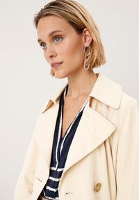 Vaaleanbeige trench-coat, jossa on läpät, kaksiriviset napit ja sileä, kevyt pinta. Yhdistetty tummansiniseen raidalliseen puseroon.