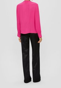 En person står klädd i en livfull rosa blus och svarta byxor, sedd bakifrån mot en neutral bakgrund.