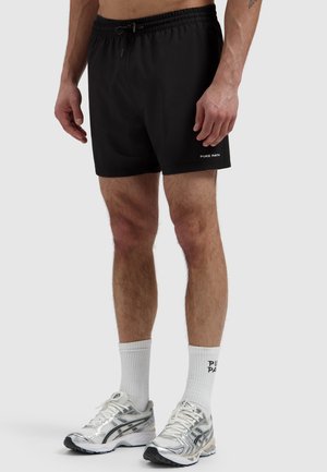 Man draagt zwarte Pure Path shorts, witte sportsokken met zwarte tekst en zilveren hardloopschoenen, staat met ontspannen armen.