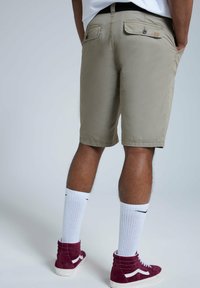 Shorts beiges avec une texture lisse, comportant deux poches arrière et une couture visible. Portés avec des chaussettes blanches et des baskets hautes bordeaux.