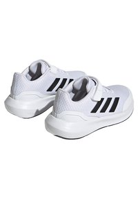 Zapatillas deportivas blancas con tejido de malla, que presentan tres franjas negras, una correa de velcro y una suela texturizada y acolchada para mayor soporte.