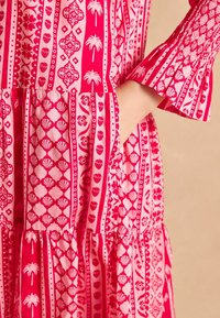 Różowa sukienka z różnorodnymi białymi wzorami, w tym muszlami i kwiatami. Materiał jest lekki, sukienka ma warstwowy krój i rozszerzane rękawy.