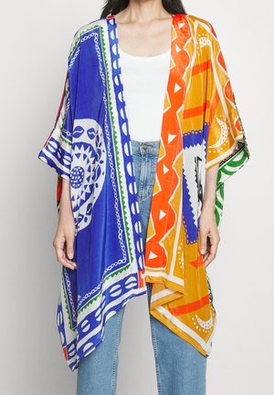 Femme portant un kimono coloré ouvert à l'avant avec des motifs géométriques et circulaires bleus, orange, blancs et verts sur un haut blanc et un jean bleu.