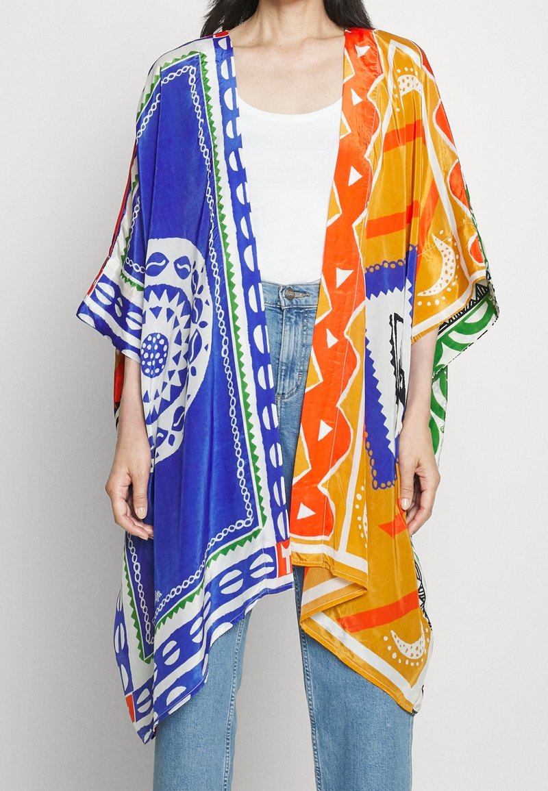 Femme portant un kimono coloré ouvert à l'avant avec des motifs géométriques et circulaires bleus, orange, blancs et verts sur un haut blanc et un jean bleu.