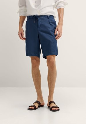 Mann trägt marineblaue Shorts mit Kordelzug, weißes hochgekrempeltes Langarmhemd und dunkelbraune Sandalen, steht auf einem ebenen Boden.