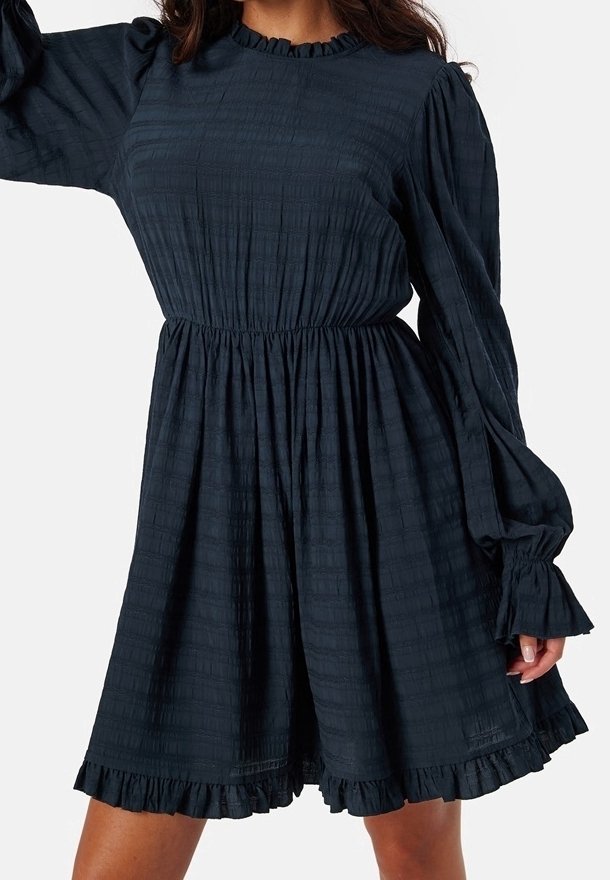 Femme portant une robe noire à manches longues avec taille élastique, col à volants, poignets et ourlet, debout devant un fond uni.