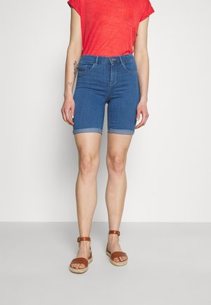 Kvinde iført opbukket jeansshorts, rød kortærmet top og brune ankelremssandaler står foran en ensfarvet baggrund.
