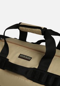 Borsa duffle beige realizzata in tessuto resistente con manici neri, dotata di chiusura con zip e patch con logo. Superficie testurizzata con impugnature rinforzate.