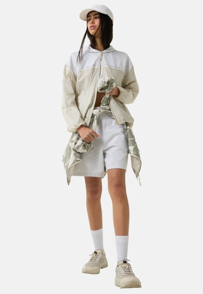 Lichtgewicht beige windjack met grijze accenten, gecombineerd met grijze shorts. Een witte pet en sneakers maken de sportieve look compleet. Opvallend camouflagedessin.